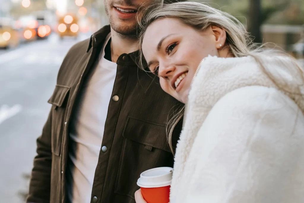 Blonde woman in a white jacket leaning on her boyfriends arm and drinking coffee outside