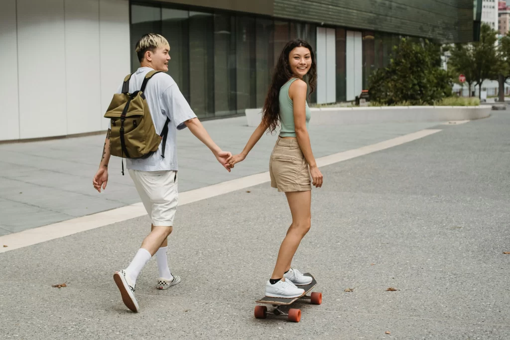 Young man and woman walking together holding hands. She is riding a skateboard