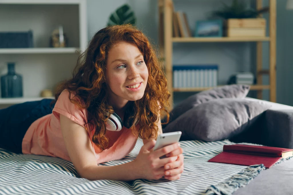 A red haired woman wearing headphones laying on her bed texting