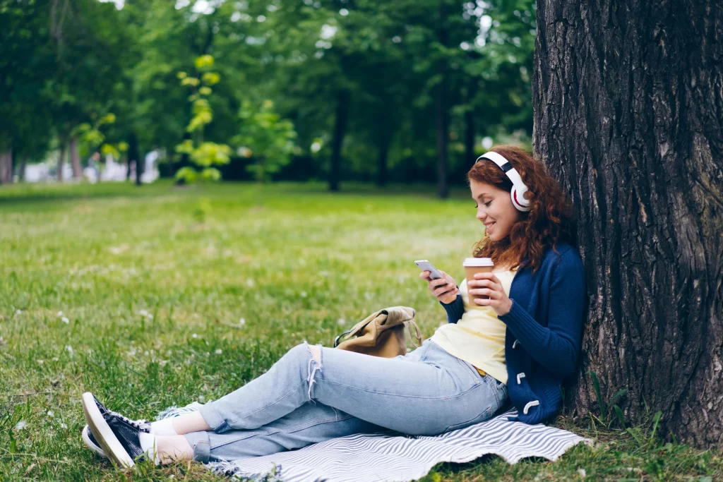 Girls with headphones in a park texting against a tree 