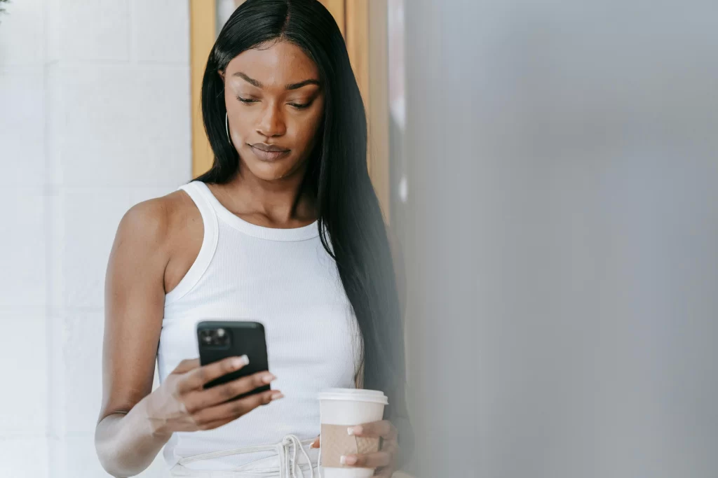 A black woman leaning on a white wall texting with a coffee in her other hand